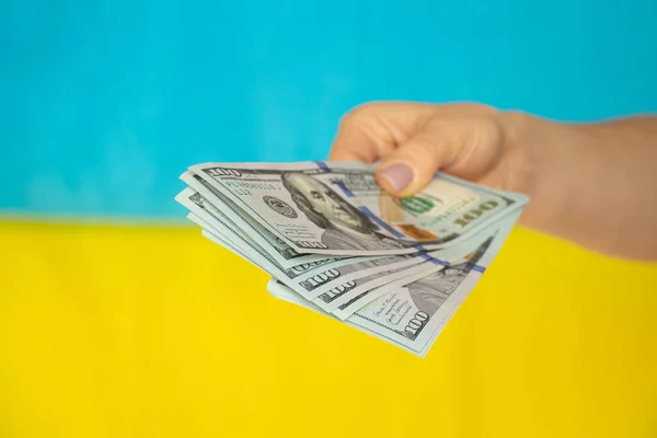 A woman\'s hand gives money in dollars against the background of the flag of Ukraine, financial support for Ukraine and money