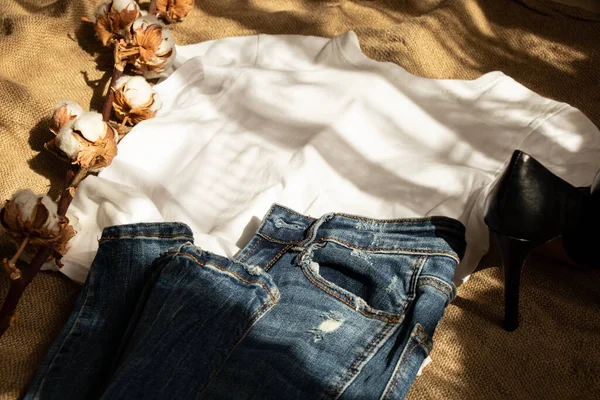 stock image White T-shirt and jeans lie on a brown burlap and a branch of cotton and shoes ,fashion clothes and shoes for women