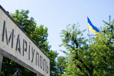 Ukrayna Mariupol 'de Ukrayna bayrağının arka planında, Ukrayna, Mariupol' de işgal edilmiş bir şehir olan Ukrayna bayrağına dayanan kurşunlarla kırılan yol tabelasının üzerindeki şehrin adı.