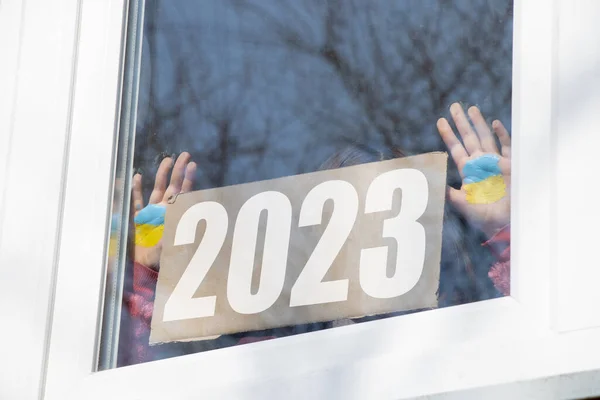 A little girl holds a cardboard with the text 2023 and the flag of Ukraine is painted on her hands in the window of her house in the Dnieper in Ukraine, the year of the war