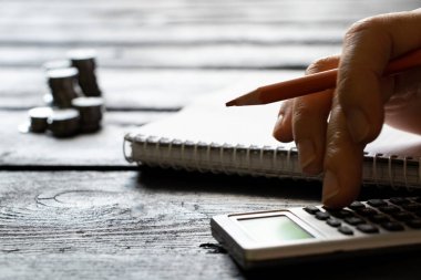 A hand on a calculator and next to it lies a notebook and stacks of coins, business and finance clipart