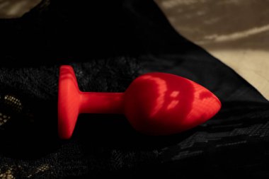 A red silicone smooth butt plug, a sex toy, lies on black lace panties in the shade. Contrast photo of anal toys, erotic toys