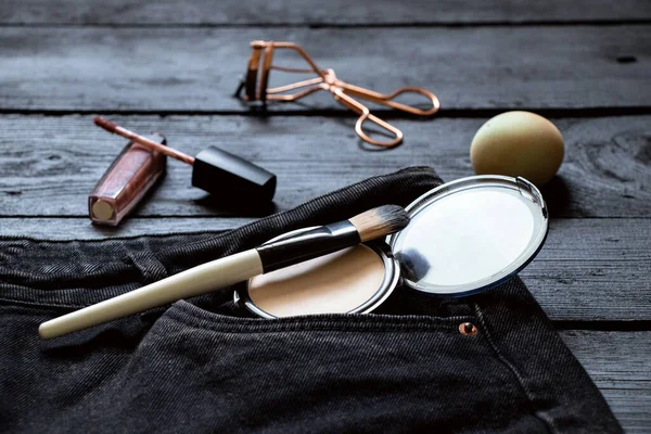 Cosmetics, powder and a brush lie in a jeans pocket, and lip gloss and a sponge lie side by side on a black wooden board,cosmetics and makeup