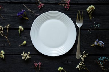 Beyaz tabak, arka plandaki dağınık çiçeklerin üzerine siyah tahta bir tahta ve yakında bir çatal, çiçekler ve yiyecek, bahar.