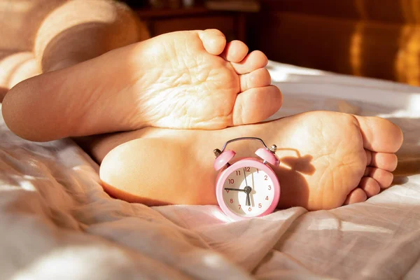 A man sleeps at home on a bed and next to a pink alarm clock, a man s legs in the morning on the bed in the sun, time to get up in the morning