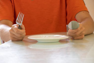 Elinde çatal, kaşık ve boş bir tabak olan bir kız evdeki mutfakta yemek ve yiyecek hiçbir şey olmadan oturuyor.