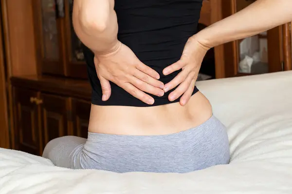 A young girl holds her back because of back pain, sitting on her bed at home, acute back pain, severe pain