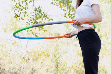 Yazın Ukrayna 'da bir parkta, belinde bir spor çemberi olan genç bir kadın bir çemberle fitness yapıyor ve fiziğini, formunu koruyor.