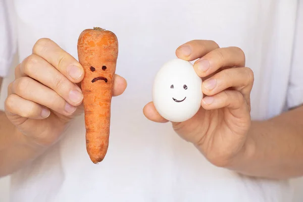 A carrot with a sad smile and an egg with a joyful smile in the hands of a girl on a white background, sadness, sadness, food products and diet