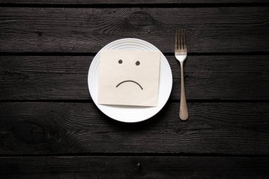 Siyah ahşap bir masanın üzerinde göğsü çizilmiş beyaz bir tabak ve kağıt yanında çatal, yiyecek bir şey yok, diyet, açlık.