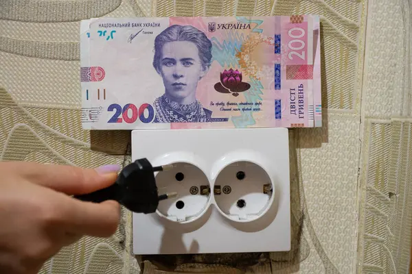 stock image A woman's hand holds a plug near a socket next to hryvnias in an apartment in Ukraine, tariff increases