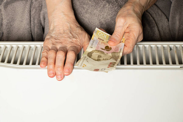 Руки пожилой женщины и украинской гривны на конвекторах в холодной квартире зимой в Украине, холодно дома, цена на тепло, высокие тарифы