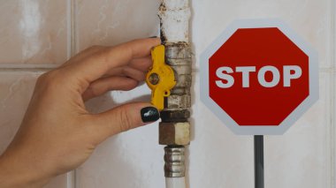 Bir kadının eli bir apartmandaki gaz borusunun musluğunu kapatıyor ve yanında dur işareti, gaz yasağı ve gaz ve yaptırımların reddi var.