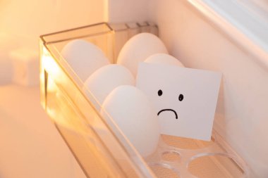 Chicken eggs are lying in a tray on a shelf in the refrigerator with a piece of paper on which a sad emotion is drawn in the kitchen, egg shortage in America
