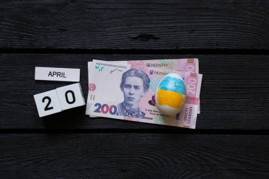 Ukrayna bayraklı Paskalya yumurtası ve 200 Hryvnia faturası ve tarihi 20 Nisan olan küpleri kara tahtada, Paskalya harcamaları, masraflar ve para