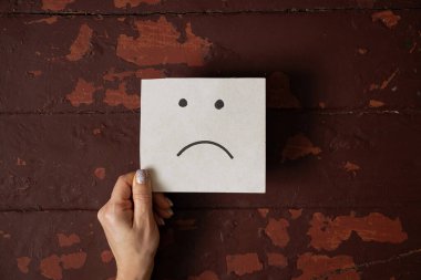 Eski bir ahşap kahverengi masanın arka planında hüzün ve hoşnutsuzluk duygusu çizilmiş bir kart tutan bir kadın.