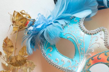 Mavi ve siyah tüylü açık mavi karnaval maskesi ve beyaz arka planda altın gül, maskeli balo için dekoratif aksesuar.