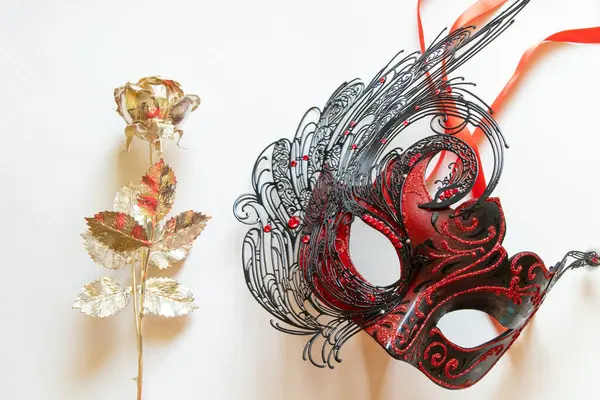 Beyaz tüylü kırmızı karnaval maskesi ve yanında beyaz arka planda altın gül, maskeli balo için dekoratif aksesuar.