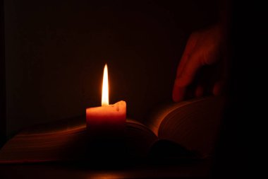Açık kitabın yanında yanan mum ve karanlığın üzerinde kadın eli, dua, İncil okuma, kitap okuma