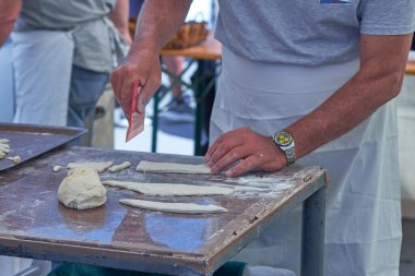 Un kaplı bir yüzeyde hamur yoğuran, önlük takan bir adamın yakın çekimi. Geleneksel hamur ve çatal bıçaklı pişirme sahnesi, sanatsal pişirme sürecini yakalıyor.