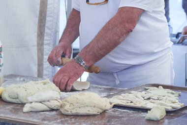 Un kaplı bir yüzeyde hamur yoğuran, önlük takan bir adamın yakın çekimi. Geleneksel hamur ve çatal bıçaklı pişirme sahnesi, sanatsal pişirme sürecini yakalıyor.