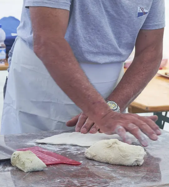 Un kaplı bir yüzeyde hamur yoğuran, önlük takan bir adamın yakın çekimi. Geleneksel hamur ve çatal bıçaklı pişirme sahnesi, sanatsal pişirme sürecini yakalıyor.