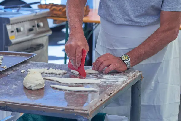 Un kaplı bir yüzeyde hamur yoğuran, önlük takan bir adamın yakın çekimi. Geleneksel hamur ve çatal bıçaklı pişirme sahnesi, sanatsal pişirme sürecini yakalıyor.