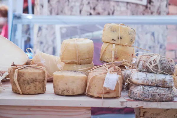 Kalabalık bir yerel pazarda farklı tipte zanaat peyniri satışa sunuluyor. Peynir doğal malzemelere sarılmış, kırsal cazibesini parlak gün ışığında vurguluyor..