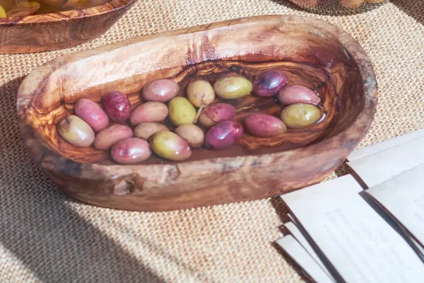 Kırsal ahşap bir kasede çeşitli zeytinler yüzer, doğal bir kumaş yüzeyine yerleştirilir. Yakınlarda, aydınlık öğleden sonra ışığında hareketli yerel pazarda ziyaretçileri bilgilendirme kartları bekliyor.