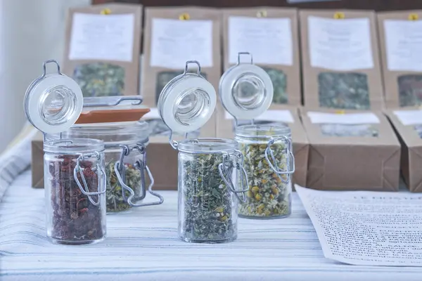 Kurutulmuş otlar ve çiçekler, bir markette masanın üstündeki cam kavanozlarda güzelce düzenlenir. Etiketler her ürün hakkında bilgi sağlar, güneşli bir günde canlı bir görüntü yaratır.