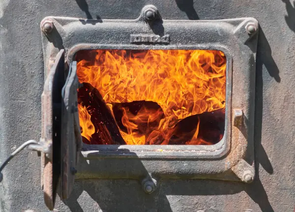 Parlak turuncu ve sarı alevlerin yakacak odun parçalarını yuttuğu, kırsal bir fırının veya ocağın dökme demir kapısının içinde kükreyen bir ateşin yakın görüntüsü..