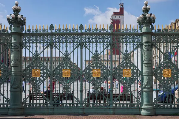 Özenle hazırlanmış, altın aksanlı demir kapılı, şehir meydanını ve gökyüzü altındaki uzak mimariyi çevreleyen ayrıntılı bir çekim..