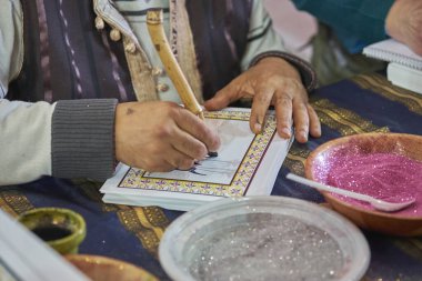 Renkli parıltılar ve desenli kumaş bir masa üzerindeki mürekkep kâseleriyle çevrili bambu kalemle geleneksel el yazısı oluşturan bir zanaatkarın yakın çekimi..