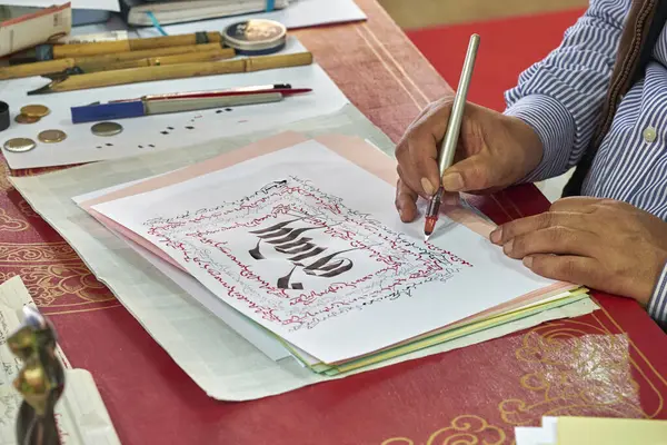 Ayrıntılı bir senaryoda kalem ve mürekkeple çalışan bir kaligrafi sanatçısının yakın çekimi. Geleneksel araçlar ve süslü kağıt kültürel miras ve sanatsal işçiliği yansıtıyor.