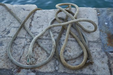 Yıpranmış uçlu deniz ipiyle sarmalanmış halat yıpranmış ucu yıpranmış eski beton iskele yüzeyinde, mavi Akdeniz liman suyunun yanındaki yıpranmış taş iskelede sakin deniz suyunun yanında düzenlenmiş halat.