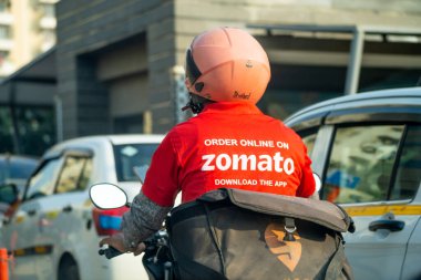 Delhi, Hindistan - takriben 2022: Hindistandaki gıda teknolojisi uygulaması ve ikonik kırmızı üniformalı Hindistan 'daki Zomato markalı sıcak kutulu bisikletçi teslimatçı çocuk.