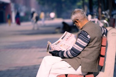 Chandigarh, Hindistan - 2023 dolaylarında yaşlı bir adam güneşli park bankında oturmuş Chandigarh 'da 17. bölgede gazete okuyor ve şehrin en ünlü alışveriş merkezini gösteriyor.