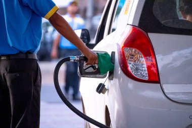 man manually filling fuel using a green nozzle into white car with people crossing showing the fuel industry in india and a major price component of Indian households clipart