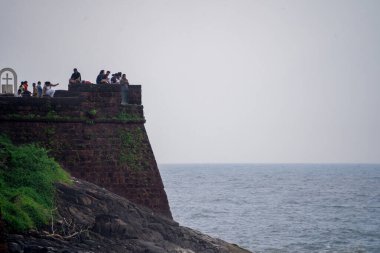 Zoomed in shot of tourists enjoying the view at Sinquerim Aguada fort with high monsoon waves at dusk splashing against the huge stone walls of the tower in goa Maharashtra India clipart