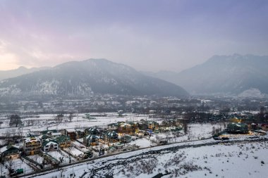 Aerial drone showing lit decorated houses in snow covered Phelgam rafting point valley with himalaya mountains and sunset sky in background in Kashmir India clipart