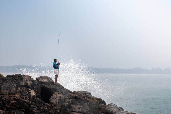 Bekal, Kerala, India - 27th Aug 2025: fisherman in lungi, dhoti, mund using a handmade fishing rod to cast a line and catch fish from the cliffs of a beach in south India
