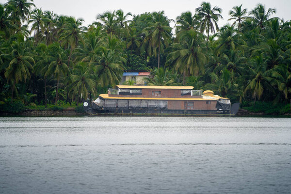 Traditional houseboat with cane bamboo roof floating away in the rain in a glassy smooth backwater in Bekal Kovalam kerela with trees in the distance showing popular tourist activity