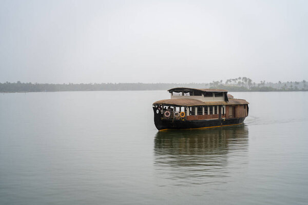 Traditional houseboat moving towards with cane bamboo roof floating away in the rain in a glassy smooth backwater in Bekal Kovalam kerela with trees in the distance showing popular tourist activity India