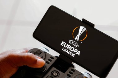 15 Ağustos 2023, Brezilya. Bu resimde, bir kişi bir oyun kumandası ile oynar ve UEFA Avrupa Ligi (UEL) logosu akıllı telefon ekranında sergilenir.
