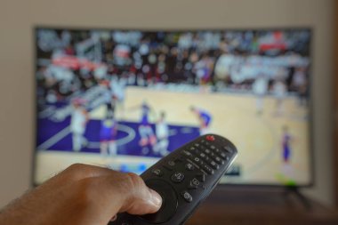 11 Mayıs 2024, Brezilya. Bu resimde, TV 'de yayınlanan bir basketbol maçının önünde uzaktan kumandayı tutan bir el var.