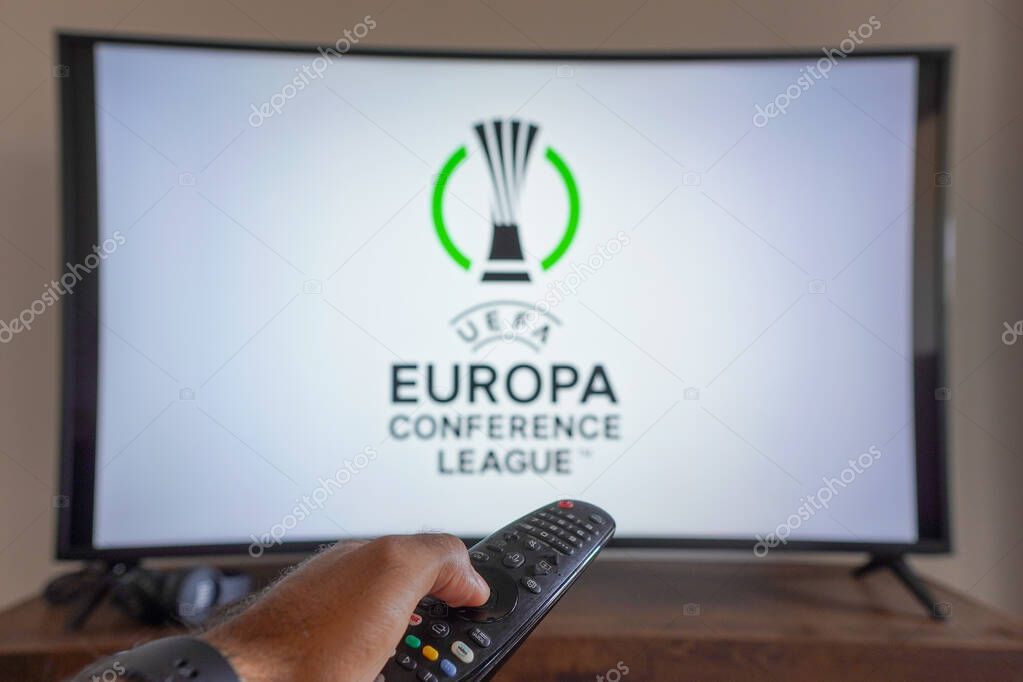 May 14, 2024, Brazil. In this photo illustration, a hand holding a TV remote control in front of the UEFA Europa Conference League (UECL) logo on a TV screen