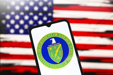 The United States Department of Energy (DOE) logo appears on the screen of a smartphone and in the background the flag of the United States on the computer screen, in Reno, United States, on December 3, 2024 clipart