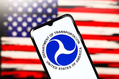ABD Ulaştırma Bakanlığı (USDOT veya DOT) logosu bir akıllı telefon ekranında ve arka planda ABD bayrağı 3 Aralık 2024 'te Reno, ABD' de bilgisayar ekranında görünür.