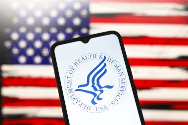 ABD Sağlık ve İnsani Hizmetler Bakanlığı (HHS) logosu bir akıllı telefon ekranında ve arka planda ABD bayrağı 3 Aralık 2024 'te Reno, ABD' de bilgisayar ekranında görünür.