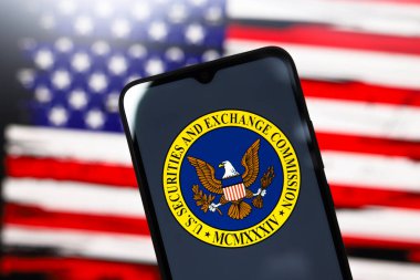 ABD Menkul Kıymetler ve Borsa Komisyonu (SEC) logosu bir akıllı telefon ekranında ve arka planda ABD bayrağı 3 Aralık 2024 'te Reno, ABD' de bilgisayar ekranında görünür.